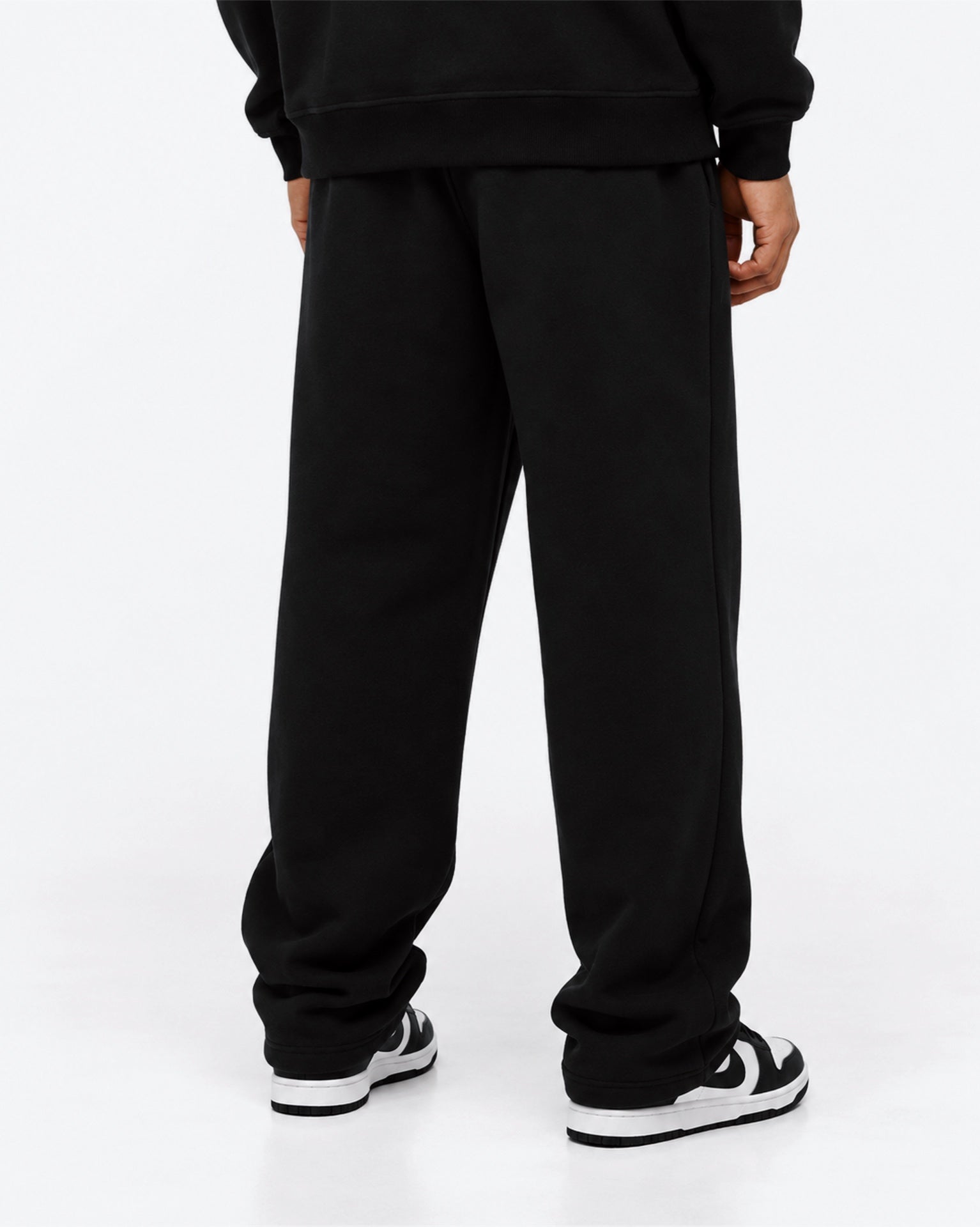 Image of A person shown from the back wearing loose black sweatpants, a black sweatshirt, and black-and-white sneakers against a clean white studio background.