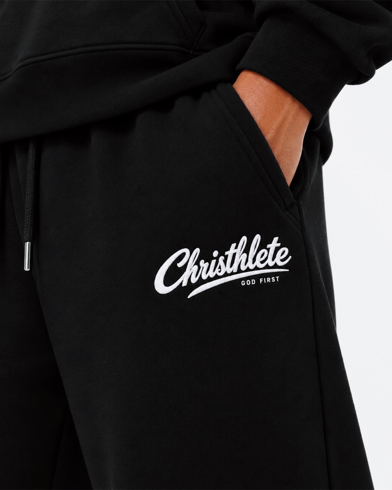 Image of Close-up of black sweatpants with a white 'Christhlete GOD FIRST' logo printed on the thigh, shown on a model with a hand in the pocket.