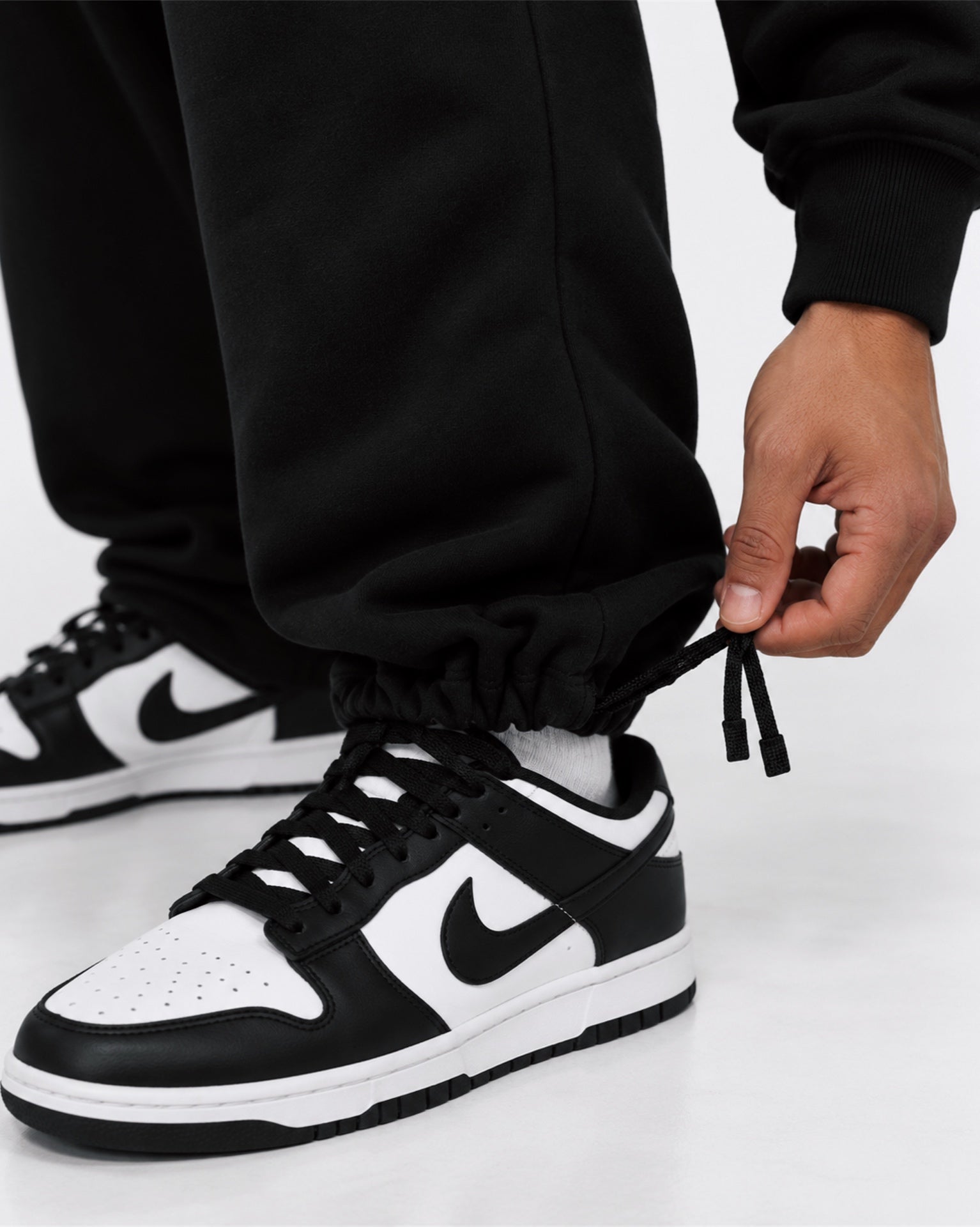 Image of A close-up of a person wearing black sweatpants and black-and-white Nike Dunk sneakers, with one hand pulling the elastic cuff of the pants.
