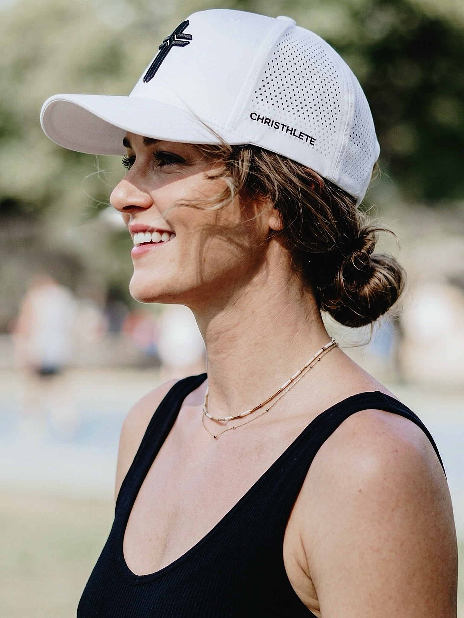 Image of A woman in a black tank top wearing a white mesh baseball cap with a black cross logo and the word 'CHRISTHLETE' on the side, photographed outdoors with a blurred background.