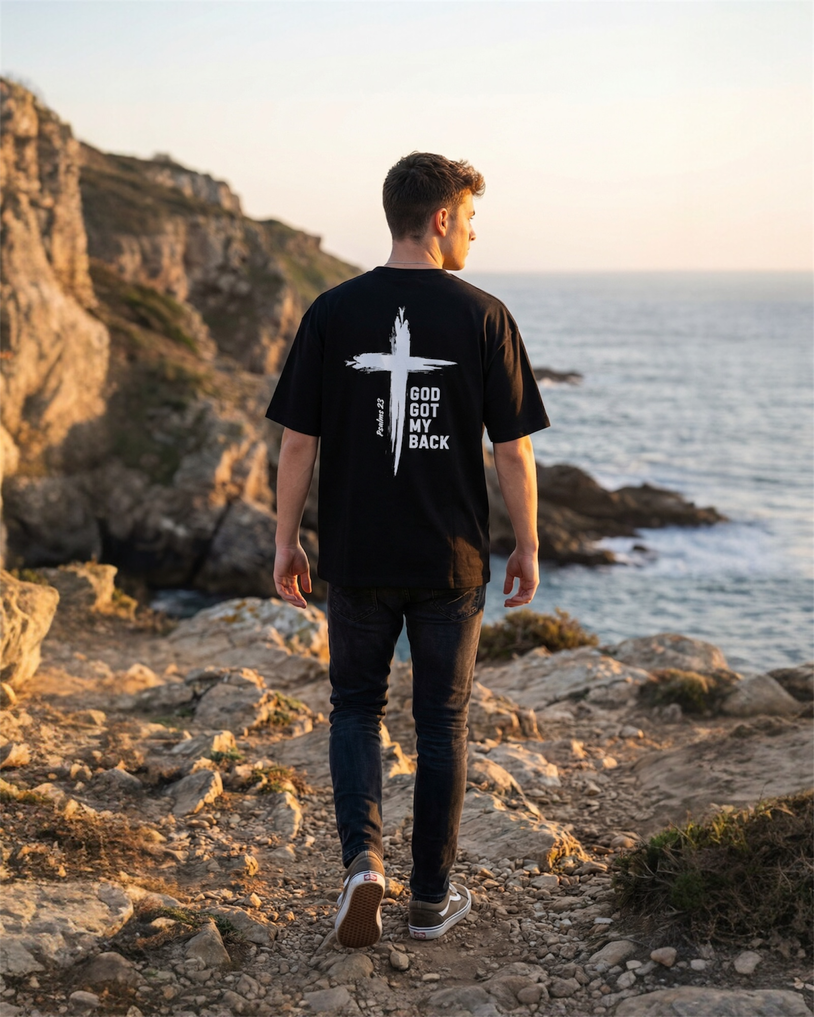 Image of Man walking on a rocky coastal path wearing a black t-shirt with a white cross design and text that says 'God Got My Back' and Psalms 23