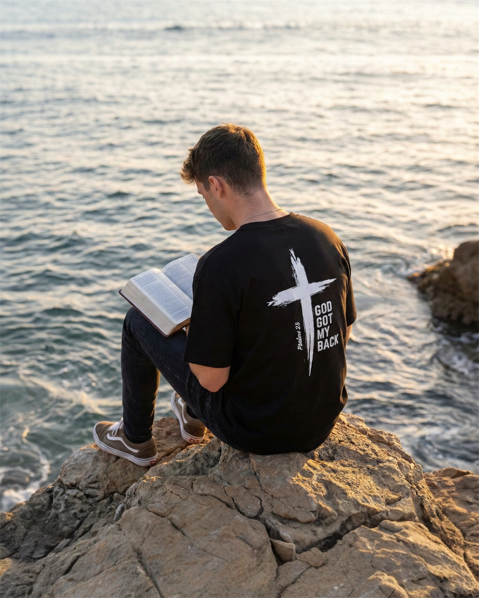 Image of Person sitting on a rock by the ocean, reading a book with a black t-shirt featuring a white cross design and text that says 'God Got My Back' and Psalms 23