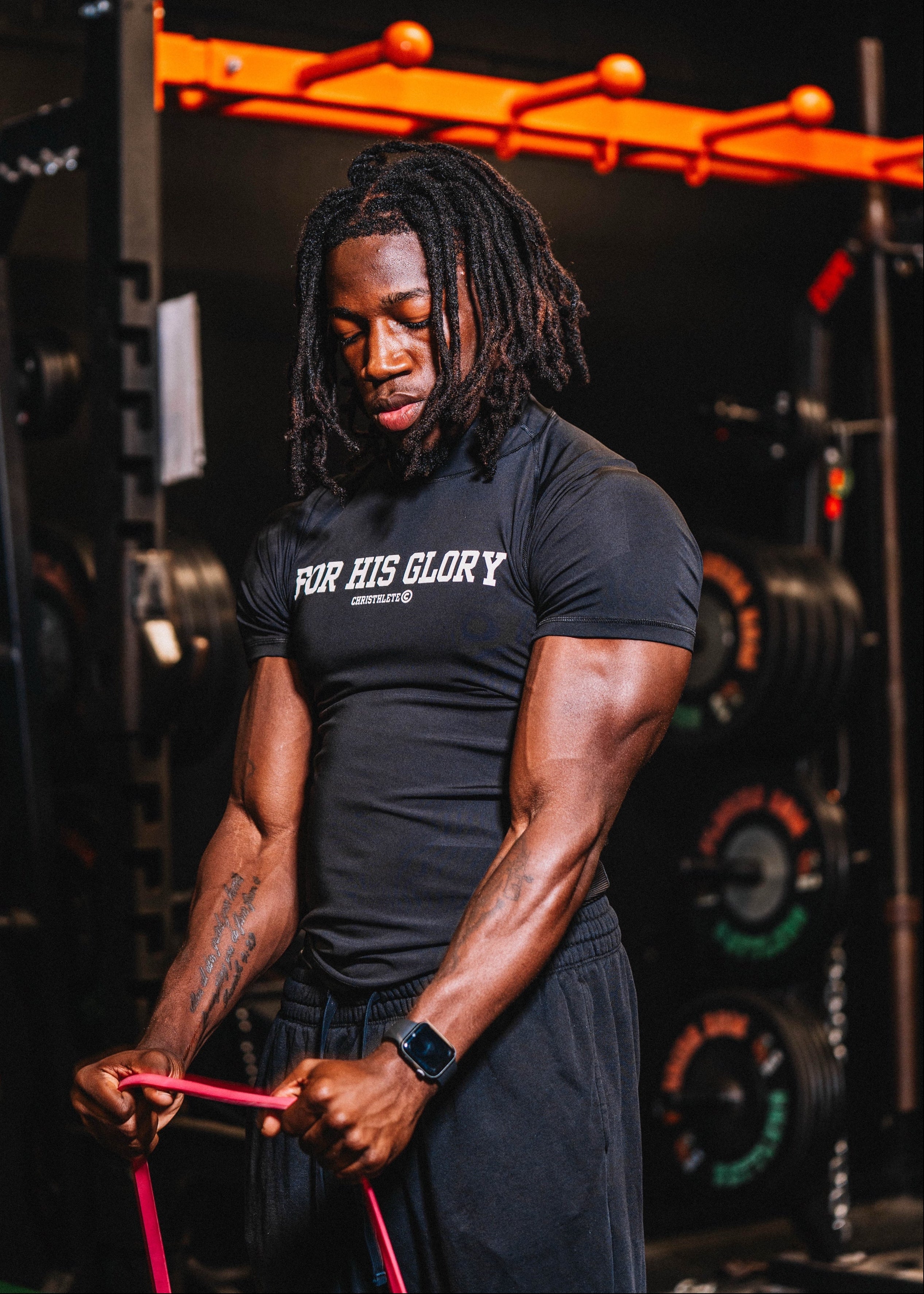 Person wearing a black 'For His Glory' compression shirt while exercising with resistance bands in a gym setting