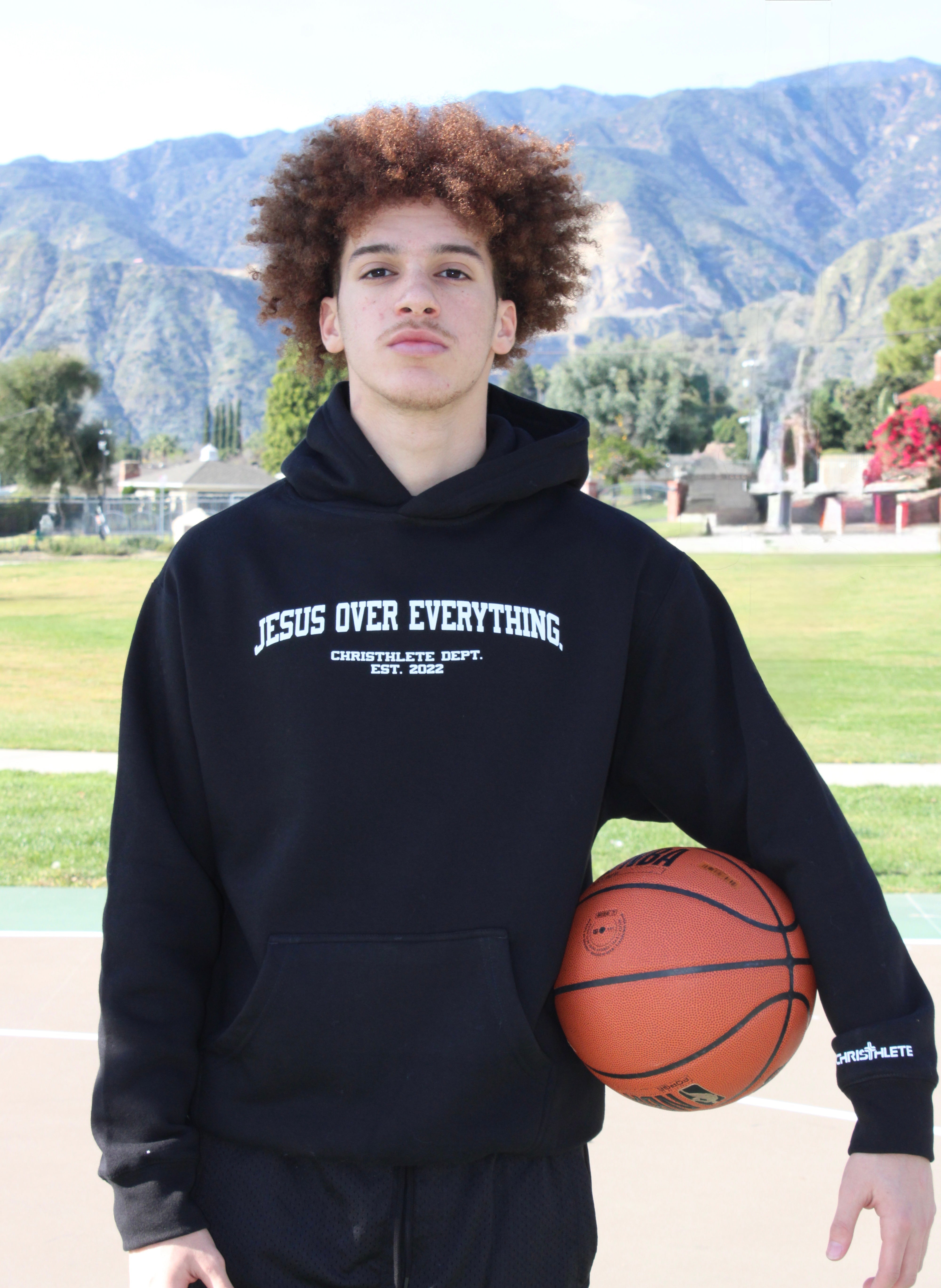Image of Athlete wearing a black hoodie with 'Jesus Over Everything' text, holding a basketball outdoors with mountains in the background.