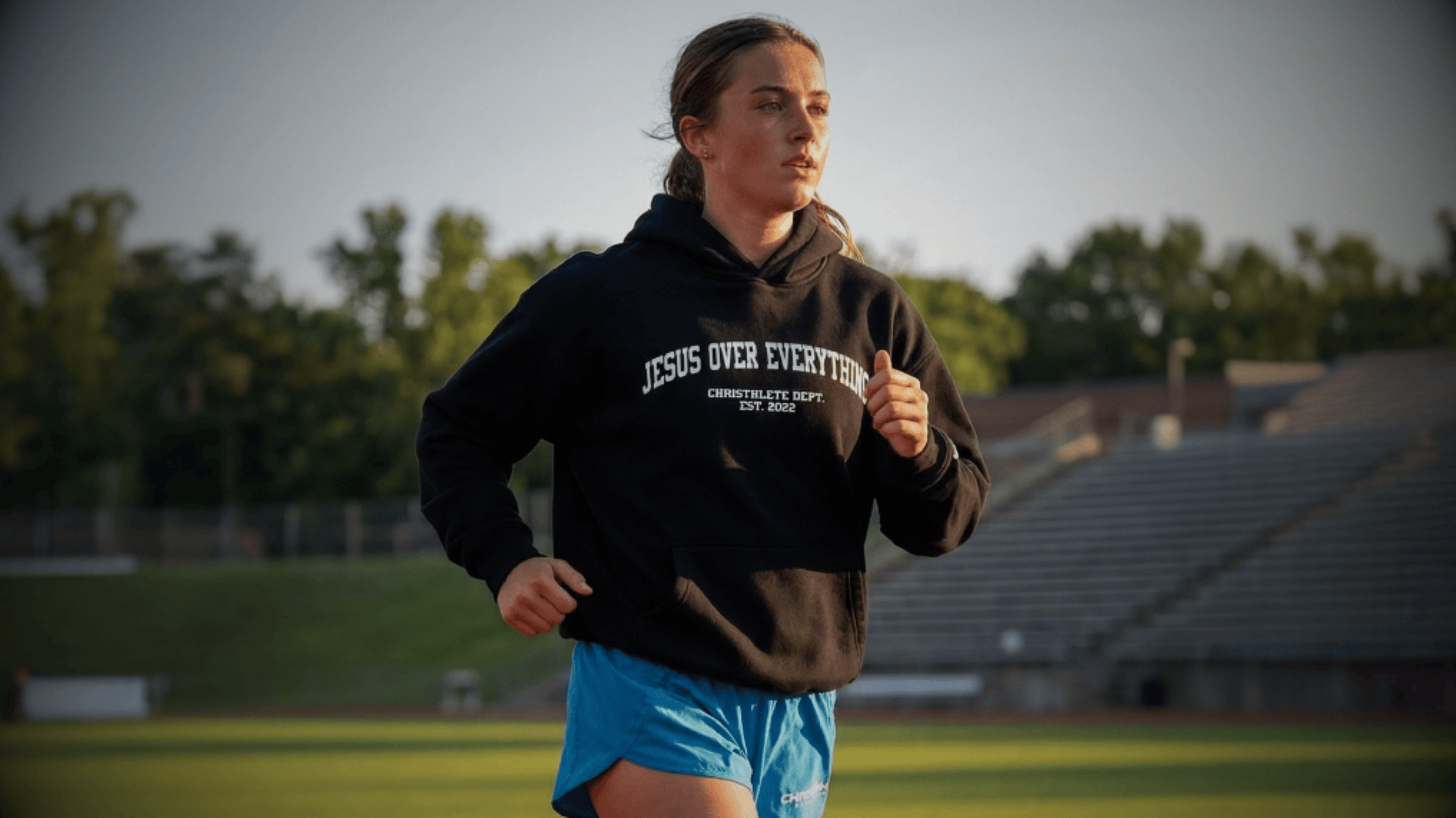 A female athlete wearing Christhlete black hoodie with 'Jesus Over Everything' text on a sports field.