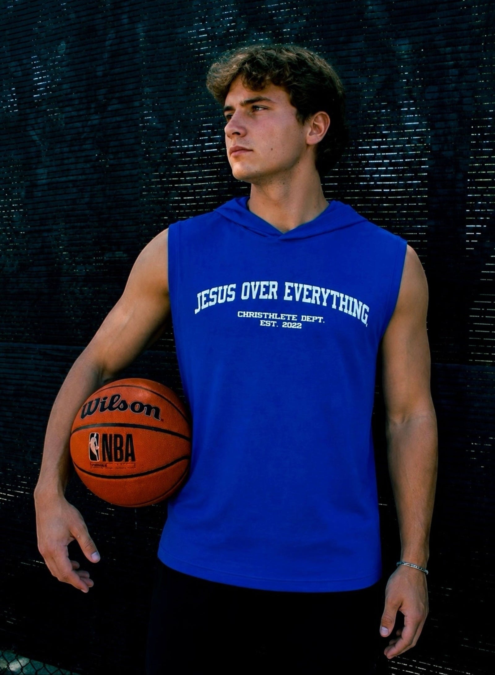 Image of Person wearing a blue 'Jesus Over Everything' sleeveless shirt, holding a basketball against a dark textured background