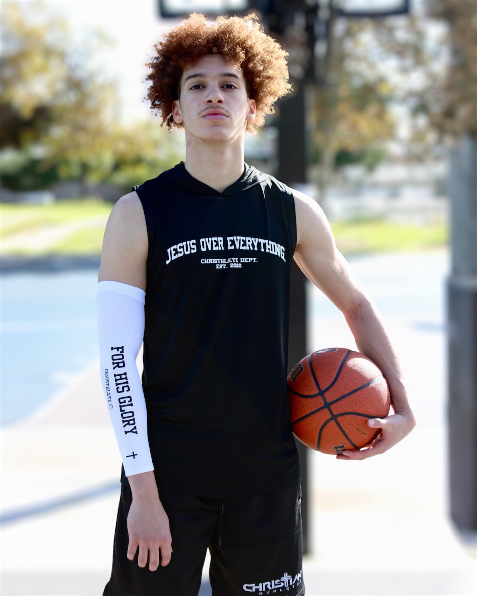 Image of Person holding a basketball wearing a black sleeveless shirt with 'JESUS OVER EVERYTHING' text outdoors.
