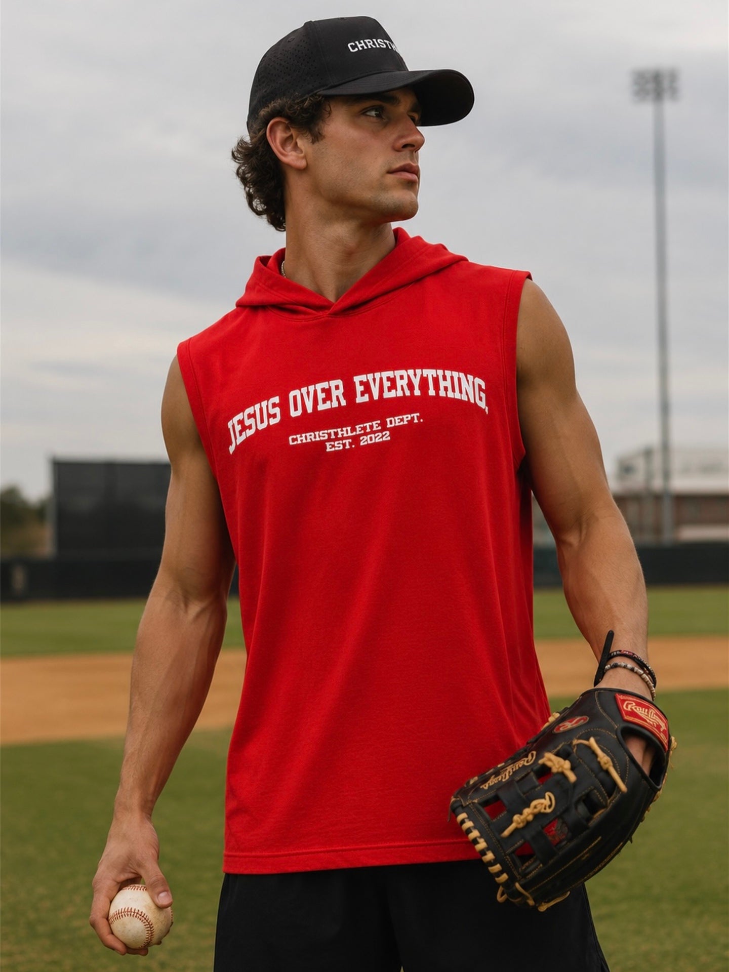 Image of An athlete on a baseball field wearing a red sleeveless hooded shirt with 'JESUS OVER EVERYTHING printed in white on the chest, a black baseball cap, and holding a baseball and a glove.