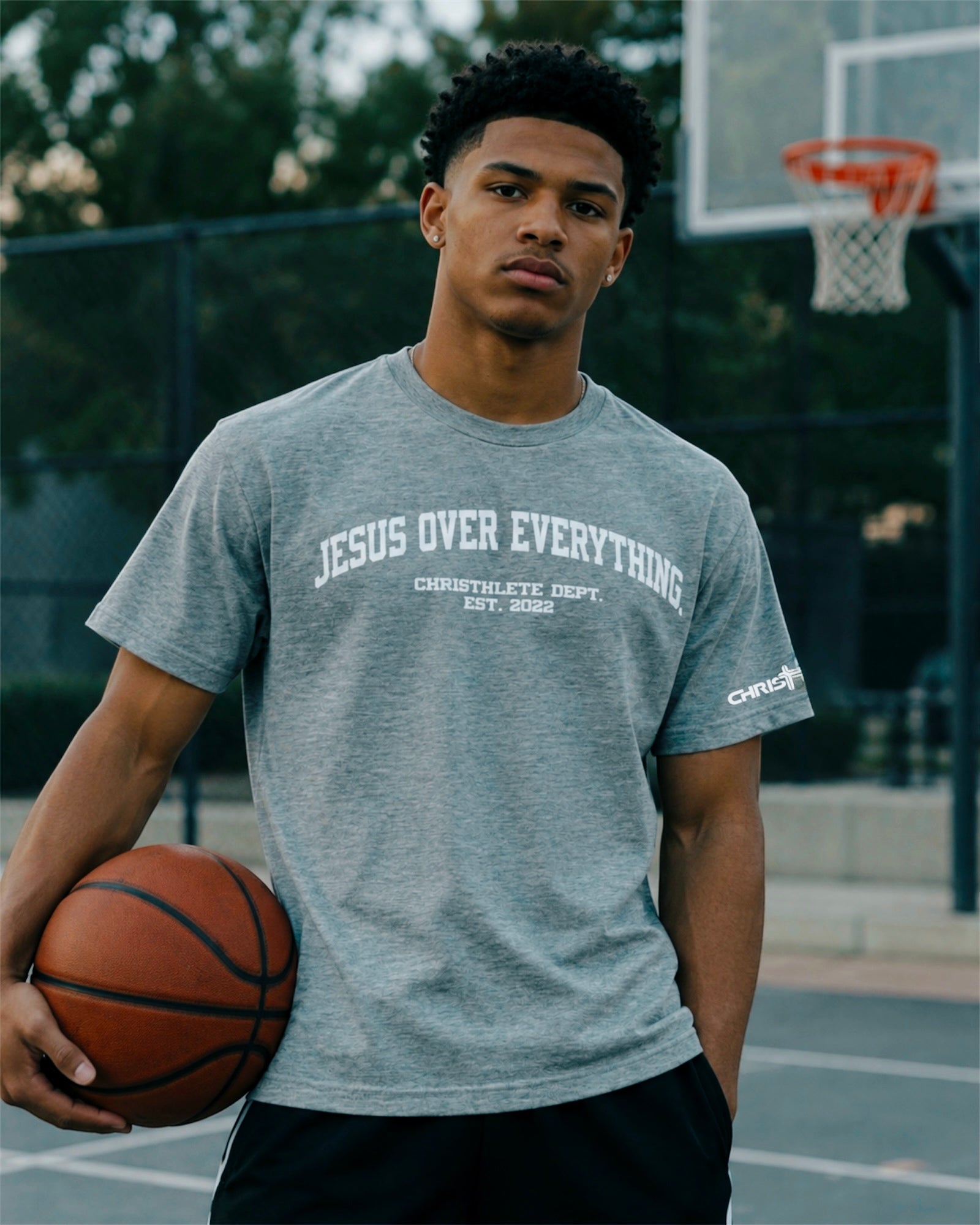 Image of A man on an outdoor basketball court wearing a heather gray t-shirt with 'JESUS OVER EVERYTHING' printed on the chest, holding a basketball.