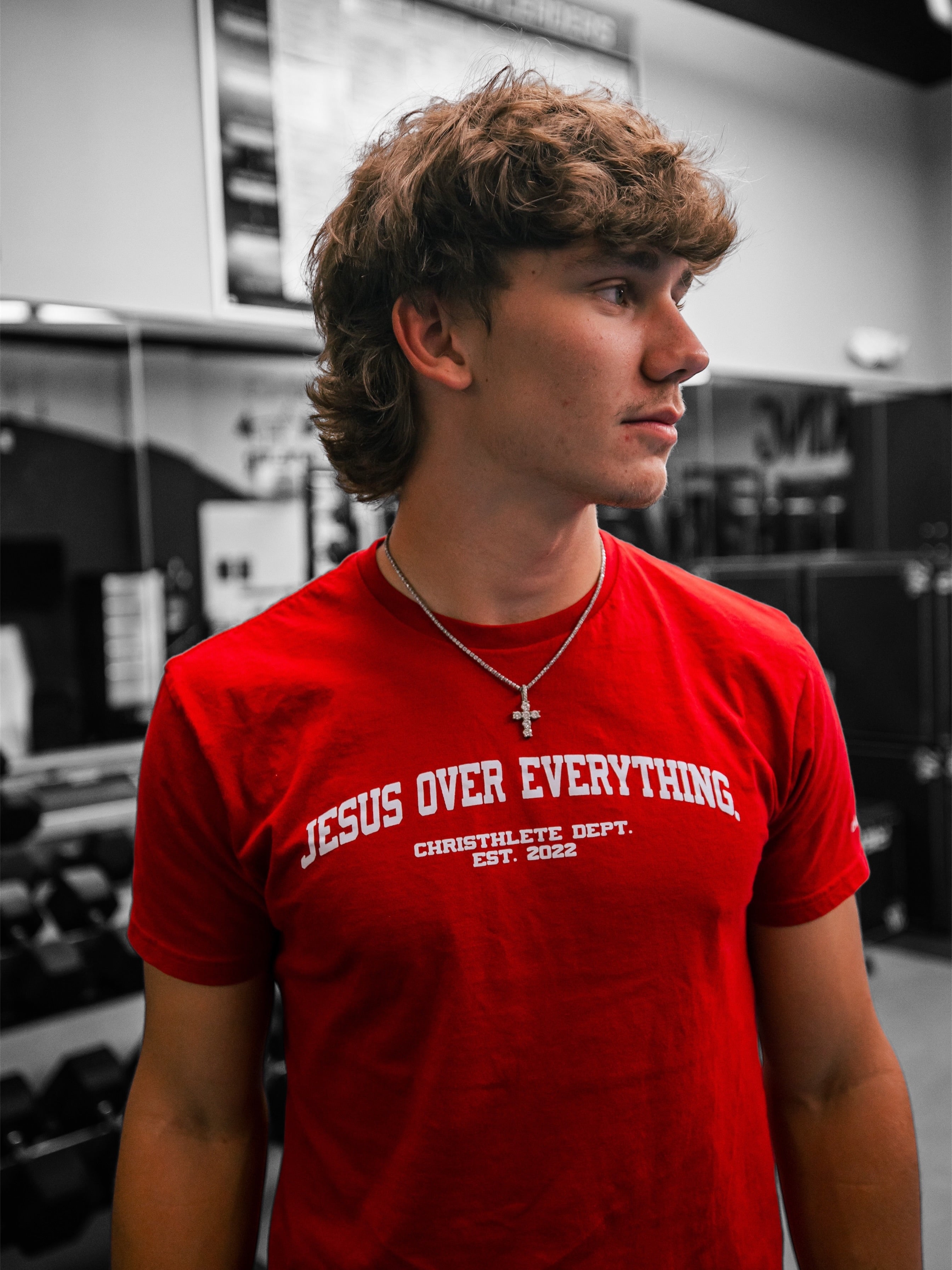 Image of A young man with curly brown hair wearing a bright red t-shirt with white text that reads 'JESUS OVER EVERYTHING' and 'CHRISTHLETE DEPT. EST.