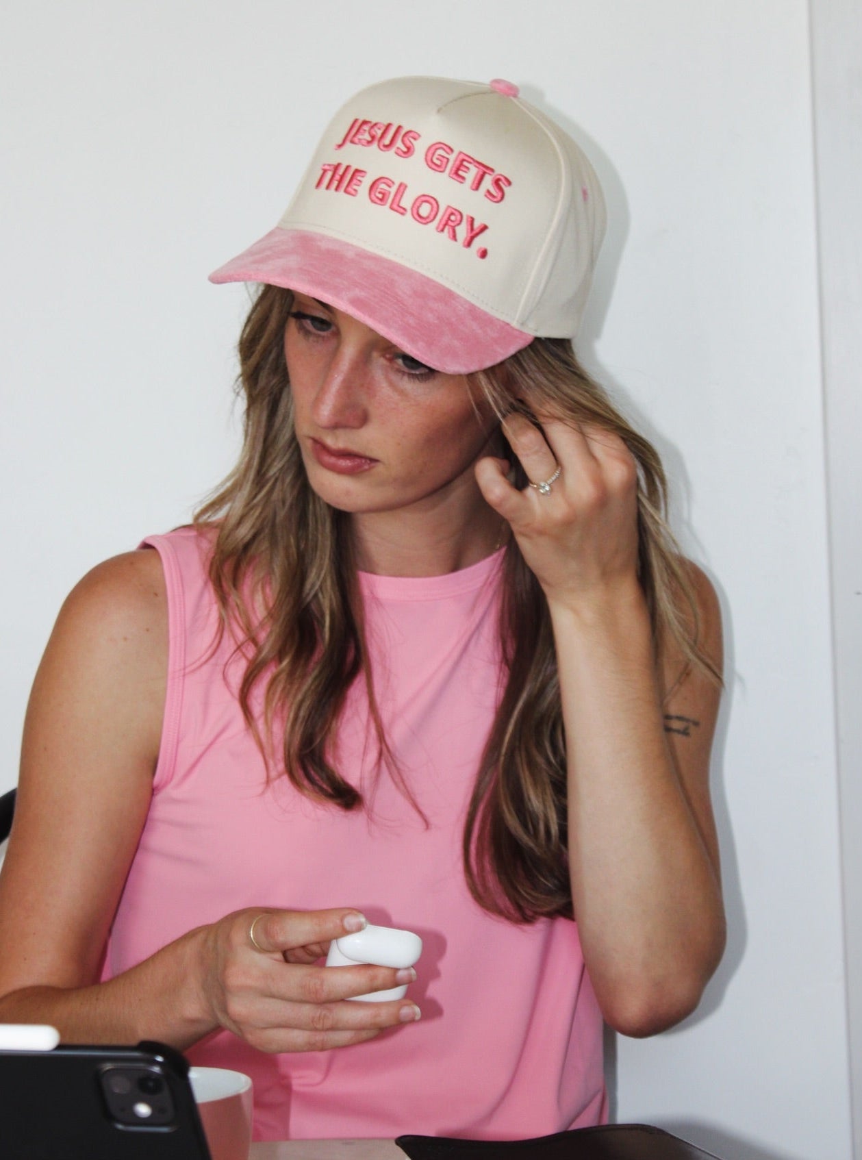 Image of A woman wearing a pink and cream trucker hat with the text 'JESUS GETS THE GLORY.' in red, paired with a pink sleeveless top, sitting at a desk with a tablet and notebook.