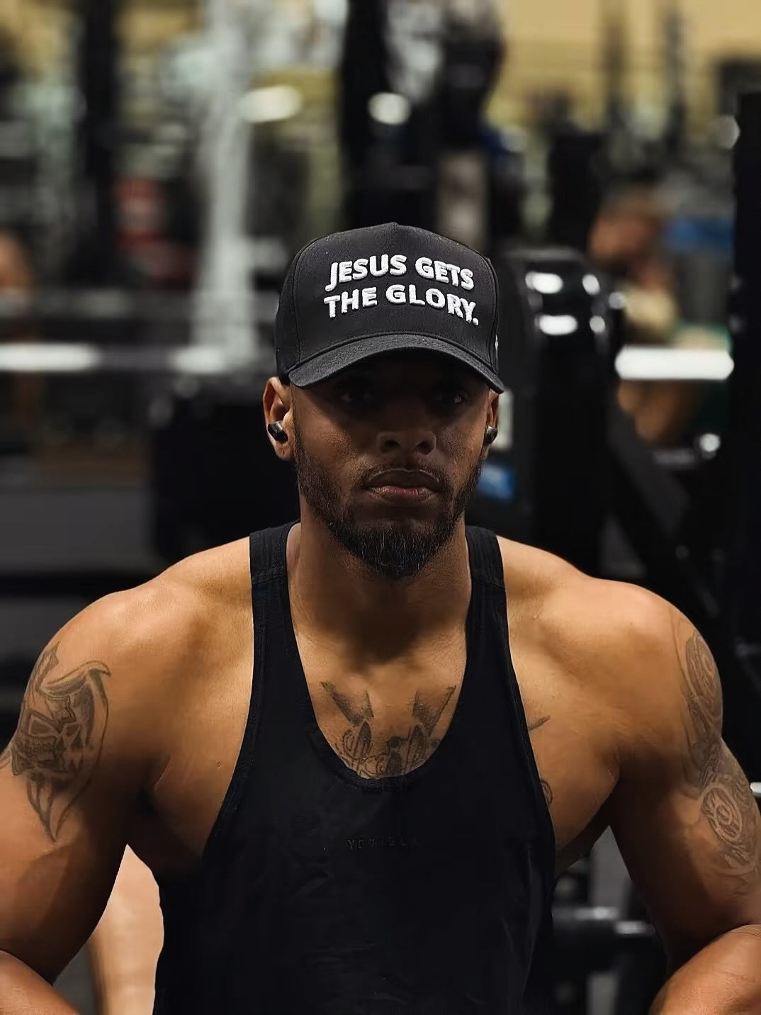 Image of A muscular man in a gym wearing a black tank top and a black baseball cap with the white text 'JESUS GETS THE GLORY.'