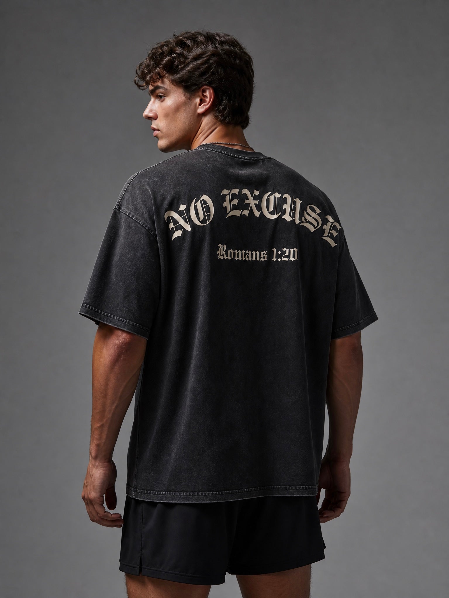 Image of A man shown from the back wearing a faded black graphic t-shirt with 'NO EXCUSE Romans 1:20' printed in a light beige gothic font, paired with black shorts against a dark gray studio background.