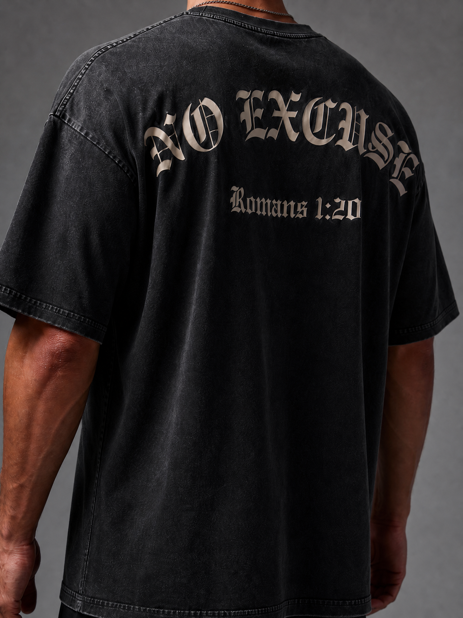 Image of A person wearing an oversized black washed t-shirt, viewed from the back. The shirt features the text 'NO EXCUSE' in a gothic font and 'Romans 1:20' underneath in a serif font, both in a light beige color.