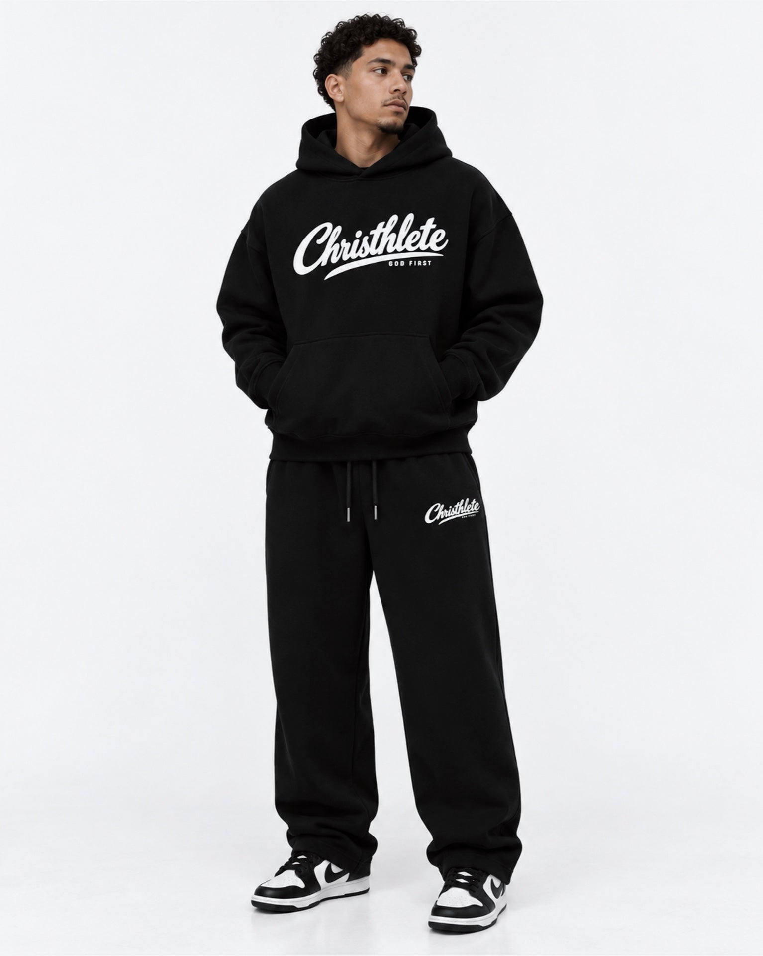 Image of A man modeling a black 'Christhlete God First' hoodie and matching sweatpants set with white logo graphics, standing against a plain white studio background.