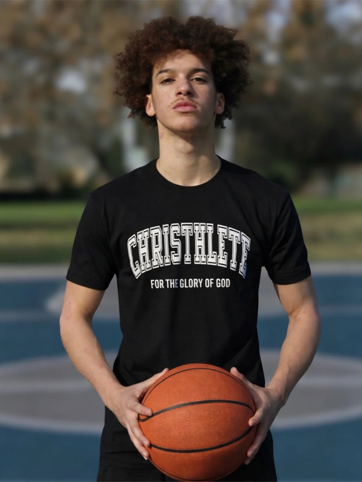 A young man with curly hair stands on an outdoor basketball court wearing a black t-shirt with 'CHRISTHLETE FOR THE GLORY OF GOD' printed in white on the front, holding a basketball.