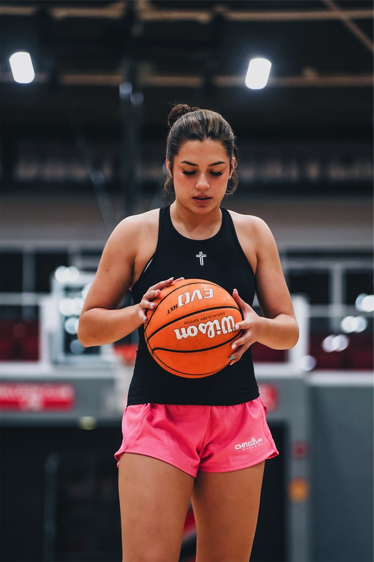 Image of Female athlete wearing a black tank top with cross logo and pink shorts with a christian athlete logo holding a basketball in a gym